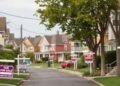 Residential street lined with houses, featuring numerous "For Sale" and "For Rent" signs on the lawns.