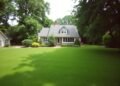 A white house with a gray roof is surrounded by a large, manicured lawn and tall trees. Another small structure is visible to the left. The scene is bright and lush.